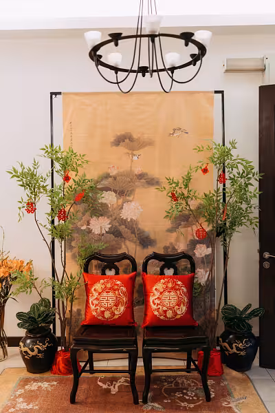 Two dark walnut chairs with red dragon and phoenix embroidered cushions before a Chinese ink painting backdrop flanked by nandina ornamental trees