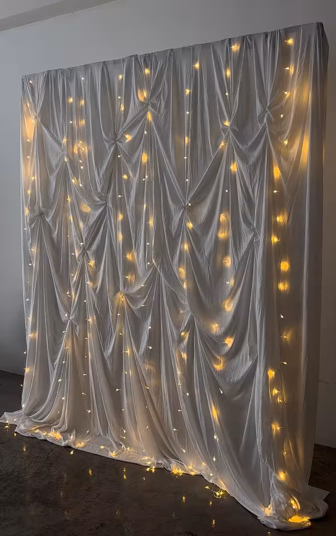 A richly ruched white fabric curtain forms a full-coverage backdrop, its gathered folds creating elegant depth and texture. Warm white LED fairy lights are layered throughout, casting a soft golden glow across the draped material for a luminous, romantic ambiance.