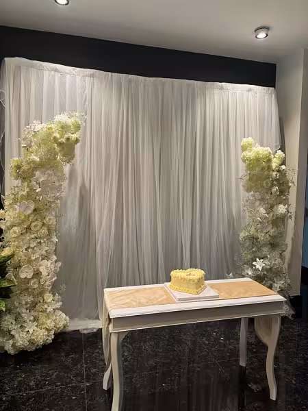 White drape backdrop framed by lush artificial flower arch columns flanking a white console table with a small cake