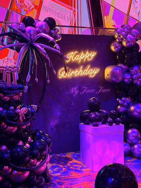 Happy Birthday LED neon sign glowing amber on a deep purple galaxy backdrop, framed by chrome purple and black balloon garlands and a sculpted balloon palm tree