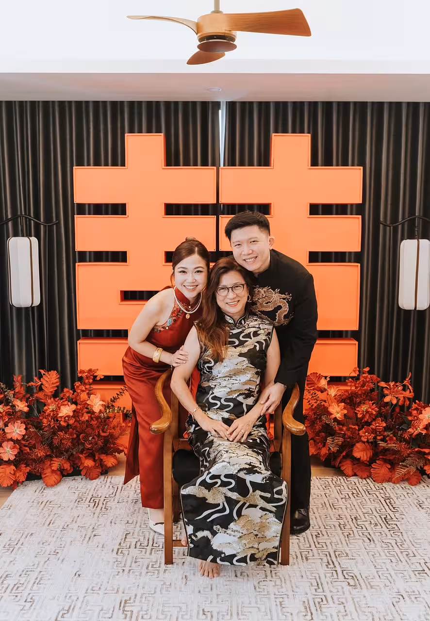Bold red 囍 Double Happiness backdrop framed by dark drapes, flanked by red artificial floor flowers and a rustic walnut wooden chair