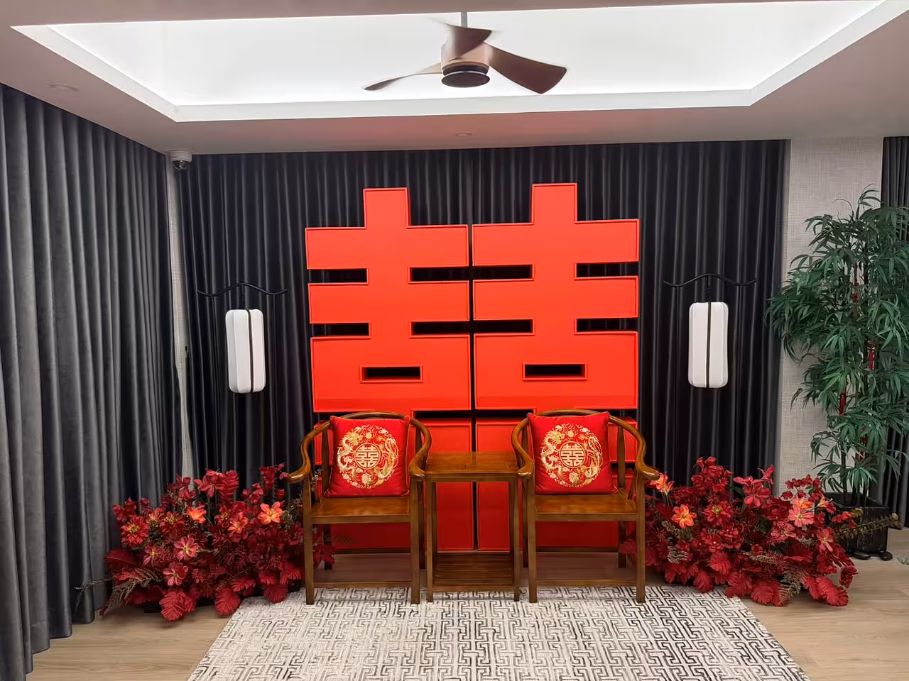 Bold red 囍 Double Happiness backdrop with walnut wooden chairs, embroidered dragon and phoenix cushions, retro arch lantern floor lamps, and lush red artificial floor flowers