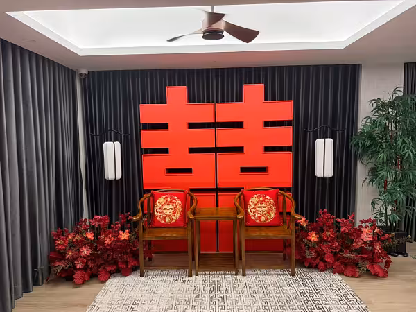 Bold red 囍 Double Happiness backdrop with walnut wooden chairs, embroidered dragon and phoenix cushions, retro arch lantern floor lamps, and lush red artificial floor flowers
