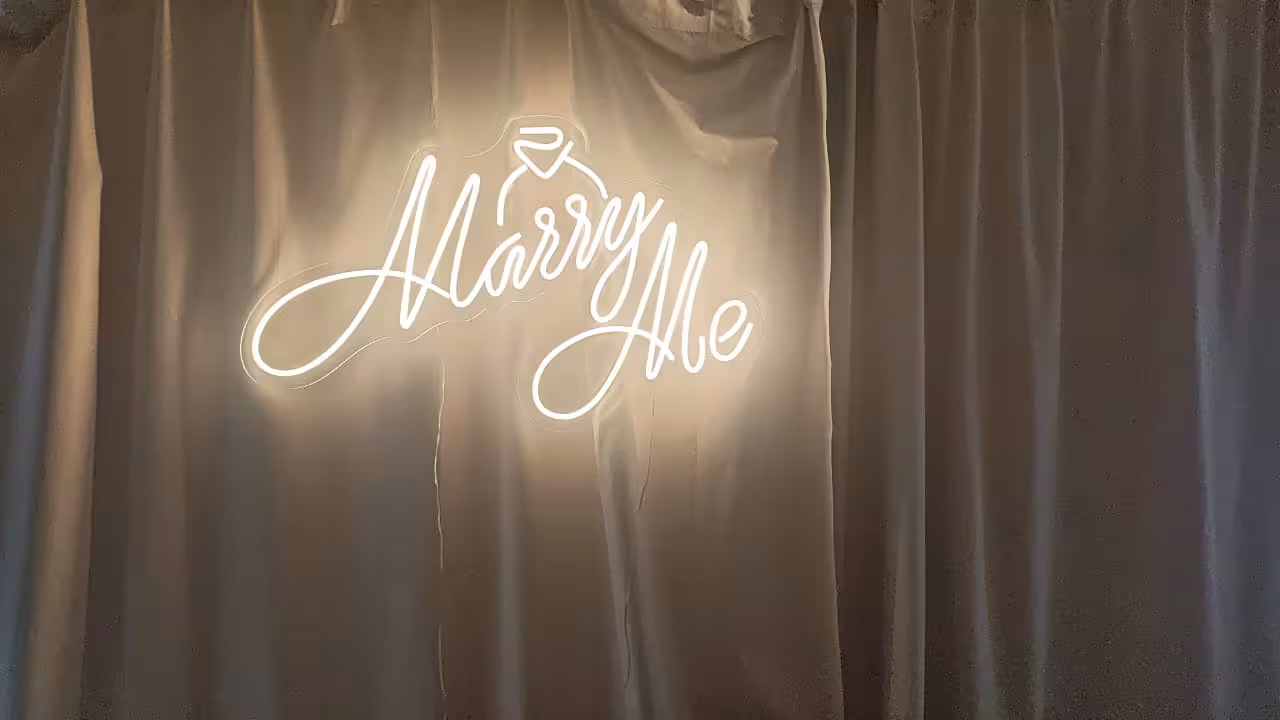 Warm white Marry Me LED neon light signage with diamond ring icon mounted on a grey satin drape backdrop