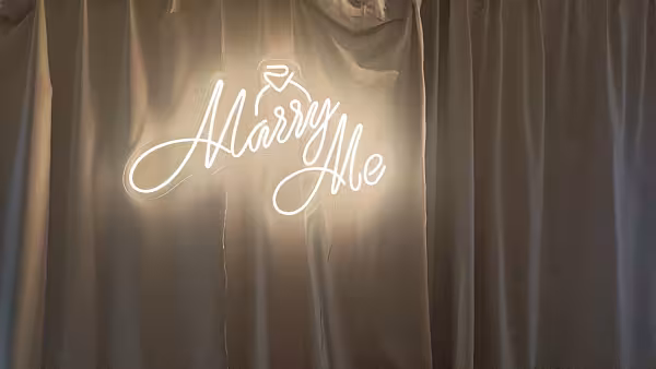 Warm white Marry Me LED neon light signage with diamond ring icon mounted on a grey satin drape backdrop