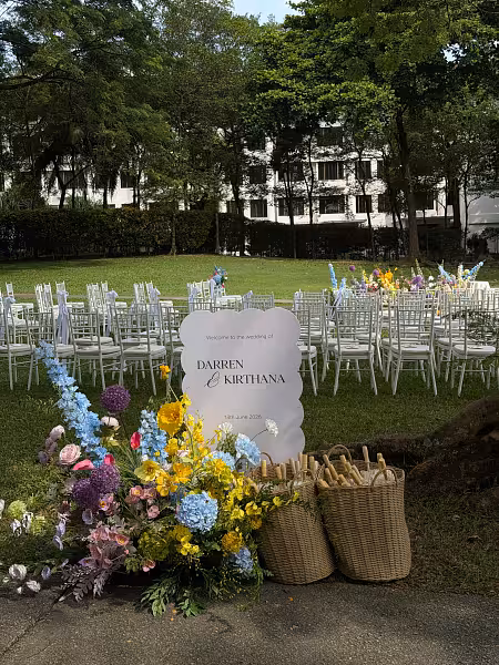 Scallop-edged welcome signboard framed by vivid blue, yellow, and pink florals, with oil paper umbrellas in woven baskets beside white chiavari chairs on a garden lawn