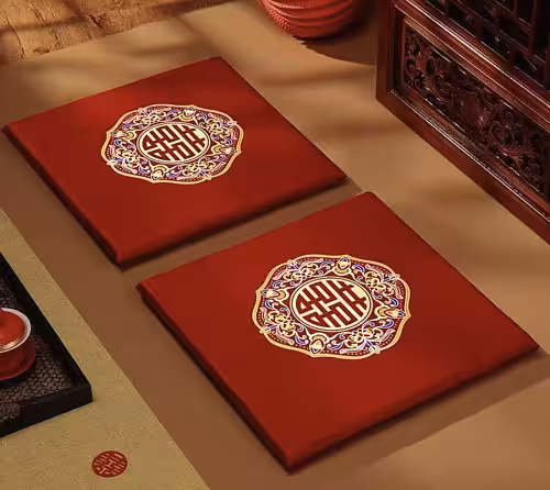 Two red satin wedding kneeling cushions laid flat side by side on a beige surface, each showing embroidered double happiness medallion, styled with carved rosewood furniture