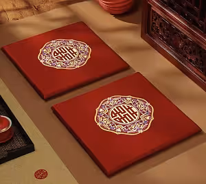 Two red satin wedding kneeling cushions laid flat side by side on a beige surface, each showing embroidered double happiness medallion, styled with carved rosewood furniture