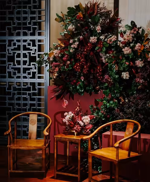 Rustic walnut wooden 3-pc chair and table set styled against a lush deep red and green floral backdrop with a Chinese lattice screen
