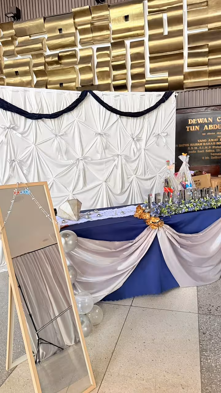 White pintuck fabric celebration curtain backdrop with navy swag trim, draped gift table with gold floral accents and silver balloons