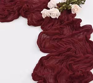 Maroon Crumpled Table Runner styled on a white surface with blush roses, showcasing its sheer, gauzy texture and ruffled drape