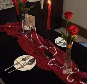 Crumpled Table Runner in maroon draped across a dark table with pearl beads, red roses, lit taper candle, and place settings in a romantic dinner setup