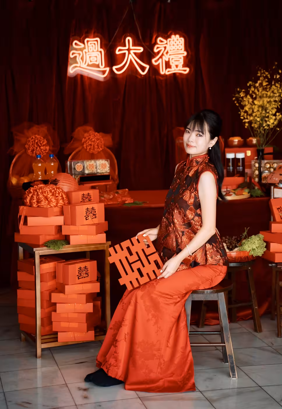 A rich wine red velvet curtain forms a dramatic backdrop, anchored above by the warm orange glow of a 過大禮 neon light. Below, stacks of traditional red double-happiness gift boxes, decorative floral accents, a paper lantern, and an arrangement of golden dried botanicals complete the auspicious Chinese betrothal display.