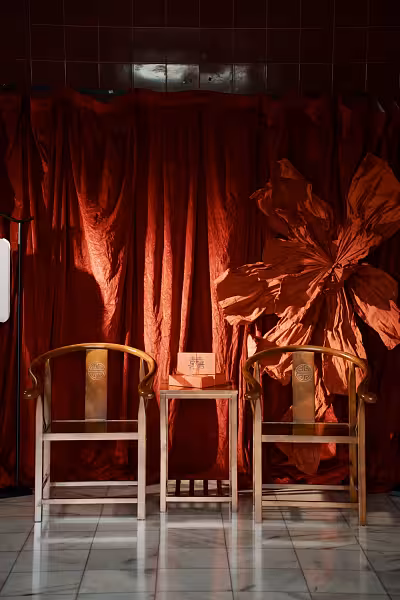 Wine red velvet drape backdrop with large red paper dielianhua flower, flanked by two rustic walnut Chinese armchairs and a centre table