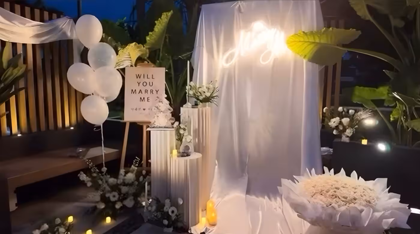 A dreamy all-white outdoor proposal setup anchors a glowing Marry Me LED neon sign against a flowing white draped backdrop, flanked by trio of origami cylindrical pleated dessert stands topped with white florals and tapered candles. A wooden easel stand displays a Will You Marry Me sign beside a white balloon cluster, while scattered tea-light candles and lush greenery complete the romantic evening atmosphere.