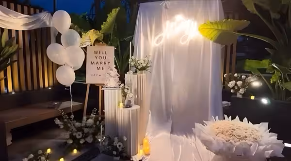 Marry Me LED neon sign on white draped backdrop with pleated cylindrical dessert stands, easel signage, white balloons, and candles outdoors at dusk