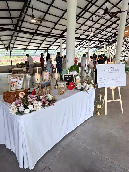 White-draped wedding welcome table with gold frames, pillar candles, blush floral clusters, and a wooden easel stand holding a wedding timeline sign