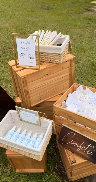Gold floating photo frames with printed signage on stacked natural wood crates, wicker baskets, and a dark wood confetti sign set on grass