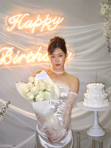 Happy Birthday LED neon sign glowing warm white on an ivory drape backdrop, with a tiered white cake on a white metal iron cake stand and white floral accents
