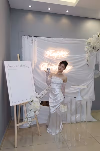 Ivory drape backdrop with glowing Happy Birthday neon sign, acrylic stands, white cake stand, LED candles, and easel signage