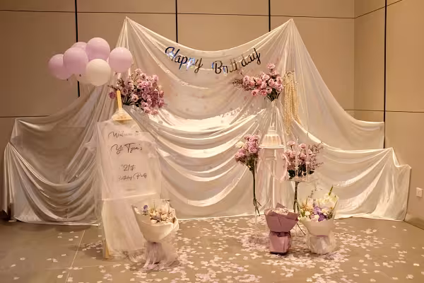 Ivory drape backdrop with mauve rose clusters, acrylic stands, easel welcome sign, pink balloons, and petal-scattered floor