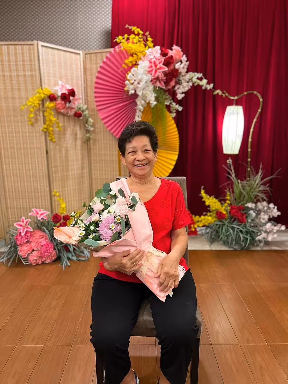 Red velvet backdrop with pink and gold origami fan arch, prosperity artificial floor flowers, bamboo screen, and antique LED floor lamp