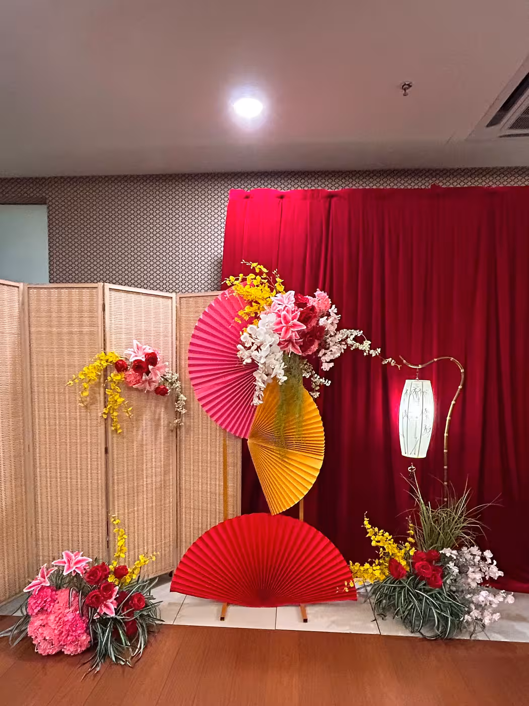 Layered red, pink, and gold origami paper fans on a wine red velvet backdrop with artificial prosperity flowers, bamboo screen, and a glowing antique bamboo floor lamp
