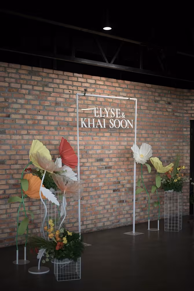 White 3m balloon flower arch with oversized paper blooms in red, yellow, orange and white, flanked by wire grid cuboid stands against a brick wall