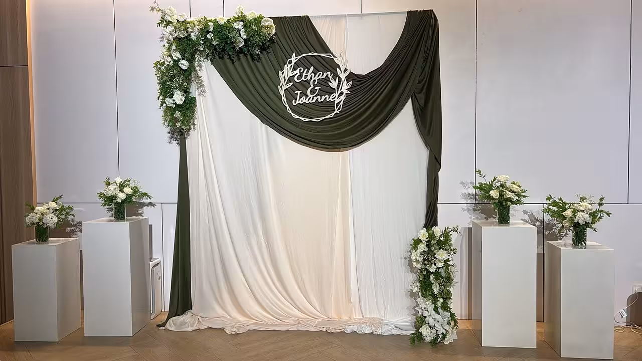 Sage green and ivory drape backdrop with white floral arrangements on cuboid pedestals and a wreath name sign