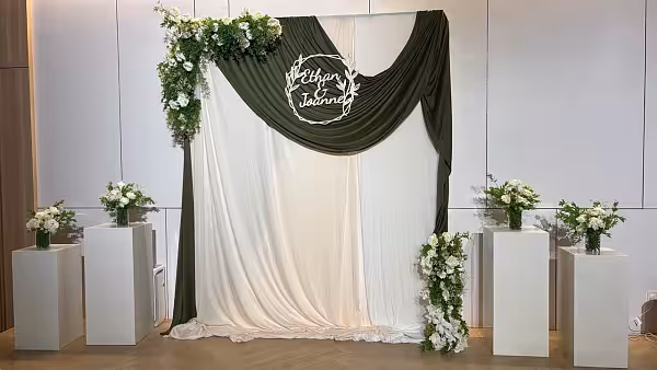 Sage green and ivory drape backdrop with white floral arrangements on cuboid pedestals and a wreath name sign