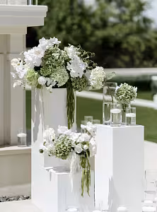 White iron cuboid dessert stands outdoors topped with white and sage green hydrangea florals, styled alongside floating candle glass cylinders