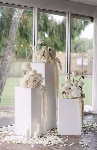 Three white iron cuboid desserts stands indoors topped with all-white rose arrangements, surrounded by pillar candles and scattered petals