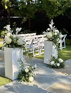 Two white iron cuboid dessert stands flanking an outdoor wedding aisle, draped with cascading greenery and white hydrangeas