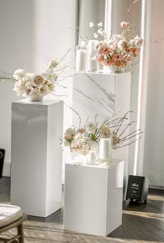 Two white glossy iron cuboid dessert stands at varying heights topped with blush pink florals and candles in a softly lit venue