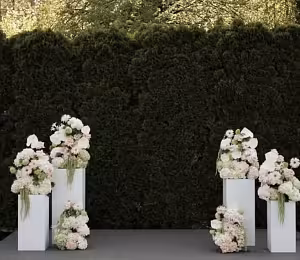 Four white iron cuboid dessert stands grouped in pairs, topped with lush pink and white floral arrangements against a dark green hedge backdrop