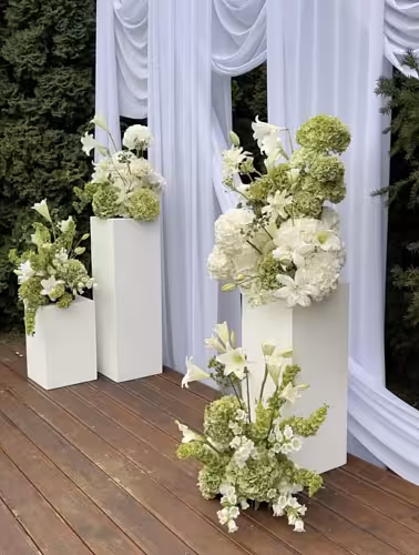 Three white iron cuboid dessert stands at staggered heights adorned with lush green and white floral arrangements on a timber deck before draped white backdrop