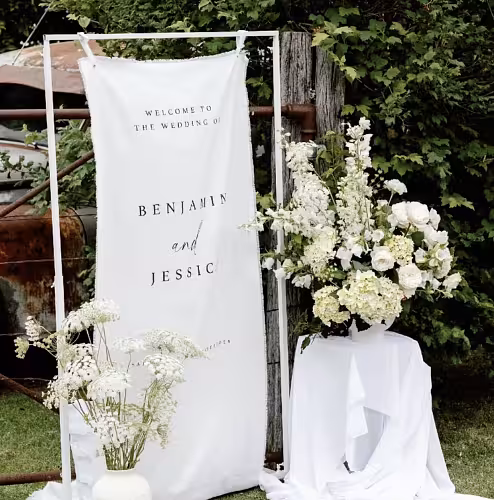 White Rectangle Arch Frame holding a draped fabric wedding welcome banner, styled with white hydrangeas and delicate wildflowers in a rustic outdoor setting