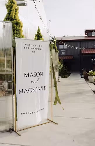 Gold Rectangle Decorative Arch Frame styled outdoors displaying a white wedding welcome banner with green ribbon accents at a farm venue