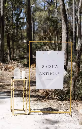 Gold Rectangle Arch Frame holding a suspended couples wedding welcome sign beside a gold cube side table in a sunlit woodland setting