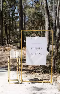 Gold Rectangle Arch Frame holding a suspended couples wedding welcome sign beside a gold cube side table in a sunlit woodland setting