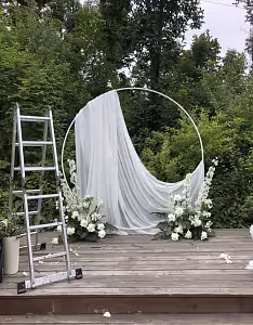White Round Stand styled outdoors with draped white fabric and white floral clusters at the base, set on a wooden deck amid lush greenery