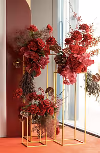 Two Gold Iron Decorative Event Stands styled with dramatic deep red roses, pink orchids, dark anthuriums, and wild branching stems against a terracotta wall