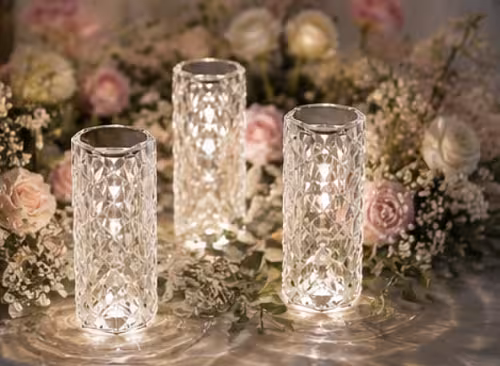 Three Crystal Rose Lamps glowing warm white together on a reflective surface, framed by pink roses and baby's breath