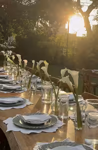 Clear glass cylinder vases holding white calla lilies styled along a wooden outdoor dining table bathed in golden sunset light