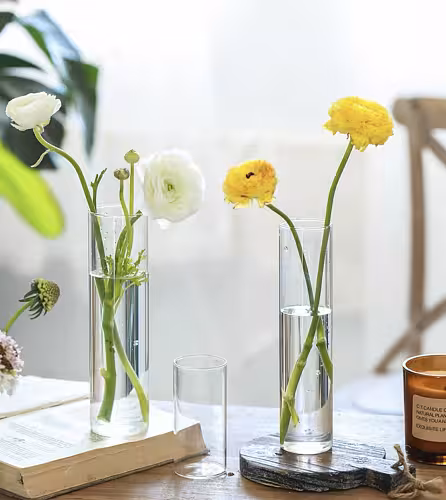 Two Minimalist Cylinder Clear Glass Vases holding white and yellow ranunculus styled on books and a rustic wooden board indoors