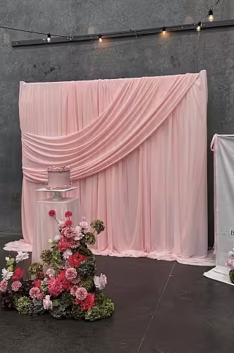 Stretchable Pink Drape Backdrop Fabric styled as a full wall backdrop with a diagonal swag gather, pink cake on pedestal, and lush floral arrangement in foreground