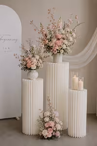 Three cream pleated cylindrical dessert stands in varying heights styled in a wedding vignette with pink florals, candles, and a J&K signage backdrop
