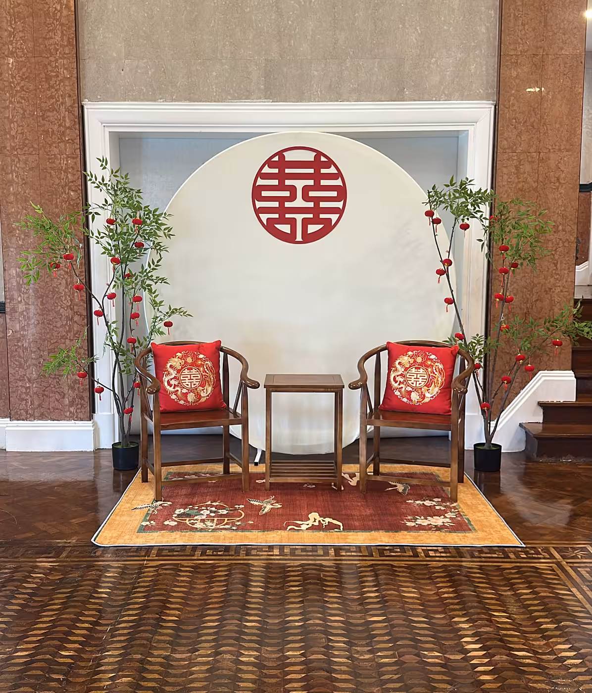 Cream circular backdrop with red double-happiness symbol, walnut chairs, dragon phoenix cushions, nandina trees with red lanterns, and floral carpet