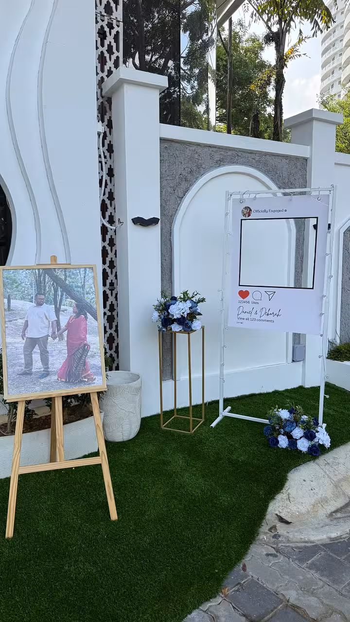 White rectangular arch frame styled as an Instagram post prop, gold iron decorative stand with navy and white florals, and a wooden easel displaying a framed photo, set on artificial grass outdoors