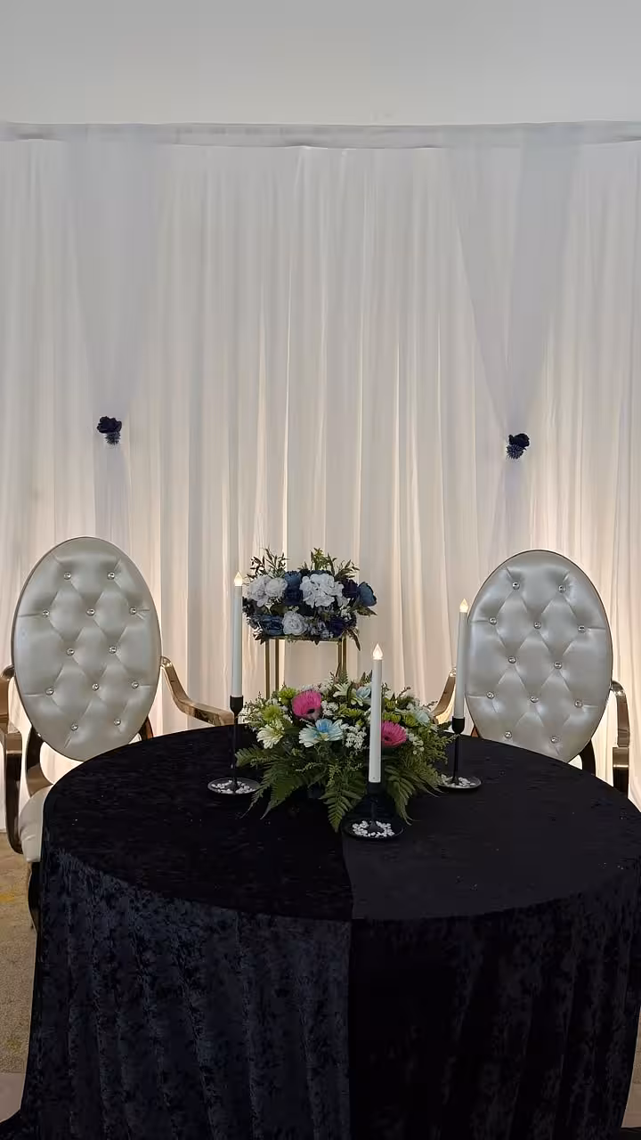 White celebration curtain backdrop with navy floral accents, black velvet round table, tufted chairs, gold iron stand, and candlesticks